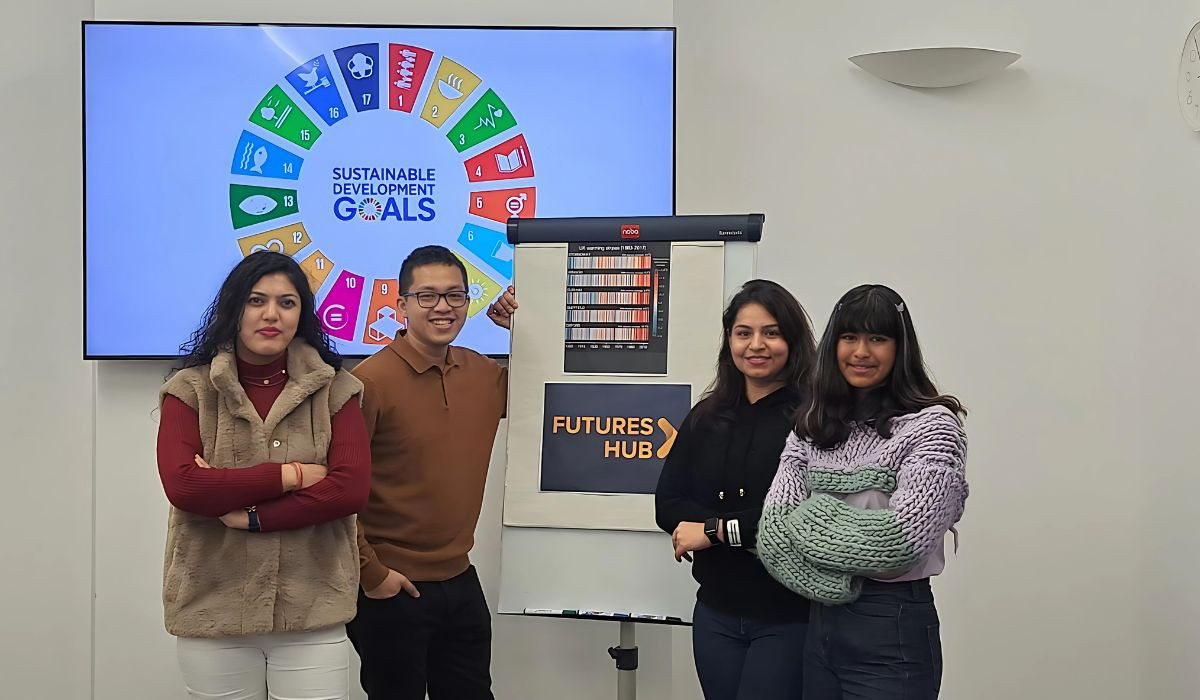 A group of four young people posing for a photo in a room with white walls, around a white board, with a sign on it that reads 'Futures Hub'. In the background there is a screen that displays the United Nations Sustainable Development Goals (SDGs). Their expressions and body language suggest they are engaged and possibly part of a team or group that is involved in projects aligned with these objectives.