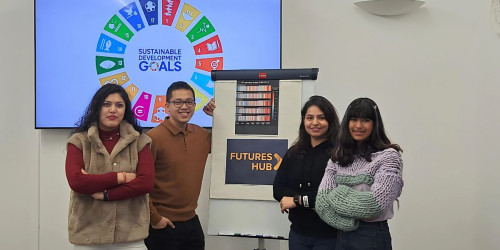 A group of four young people posing for a photo in a room with white walls, around a white board, with a sign on it that reads 'Futures Hub'. In the background there is a screen that displays the United Nations Sustainable Development Goals (SDGs). Their expressions and body language suggest they are engaged and possibly part of a team or group that is involved in projects aligned with these objectives.