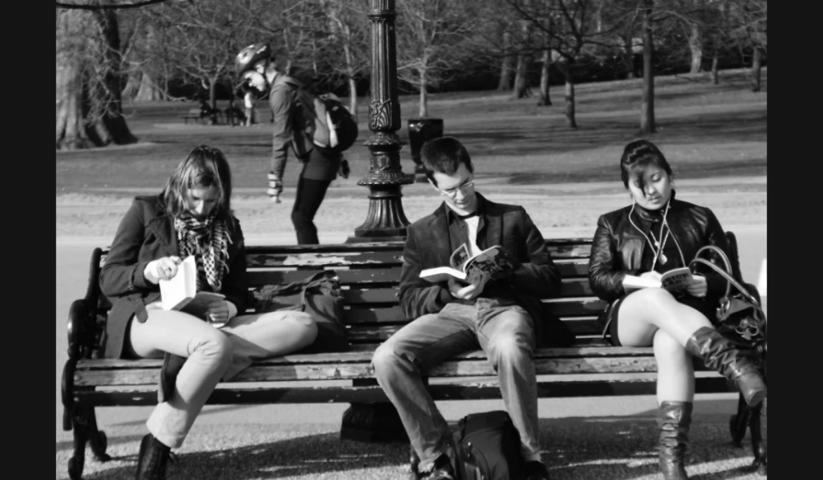 Black and white image of three people (two women, one man)sitting on a bench in a park. A person wearing a helmet is skating in the background