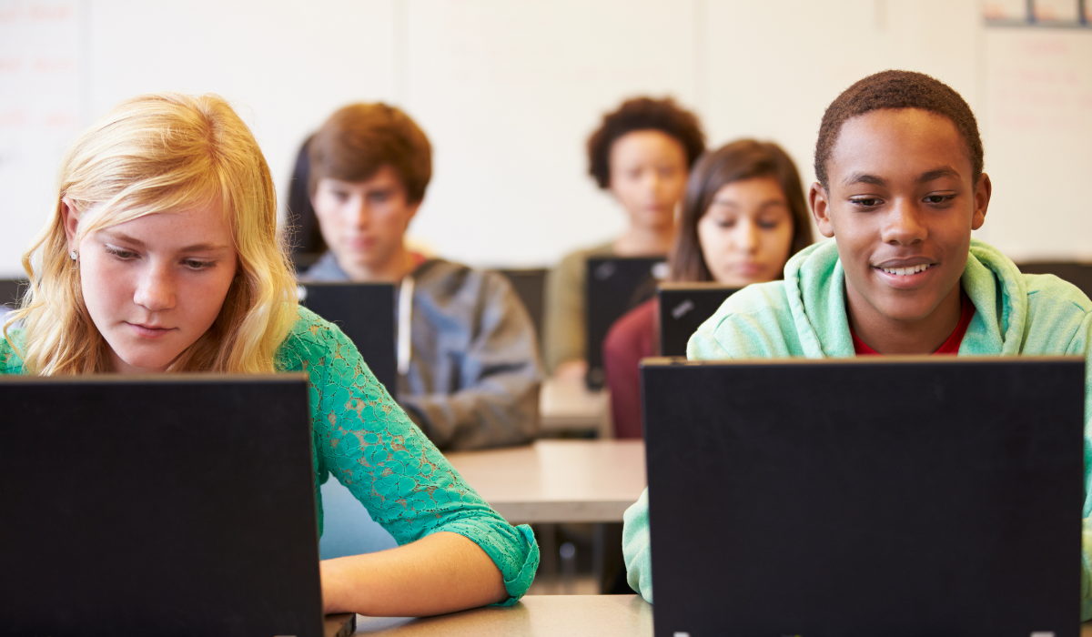 A group of students in a classroom, each working on a laptop. The focus is on two students in the foreground, one wearing a green top and the other in a light green hoodie, both engaged in their work.