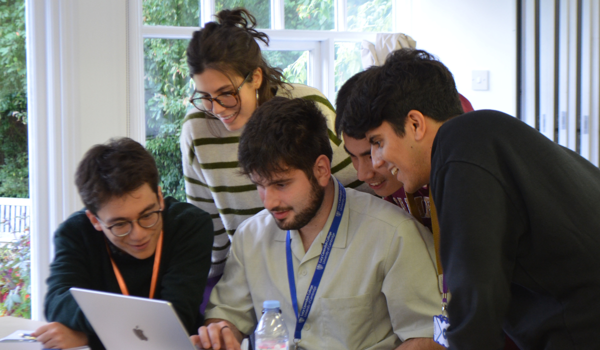 A group of five young adults gather around a laptop, engaged and smiling. They appear to be collaborating on a project, with one person seated in front of the laptop and the others leaning in, looking at the screen. The setting is the Wood-Legh room, Strathaird. There's natural light coming through large windows in the background, showing greenery outside.