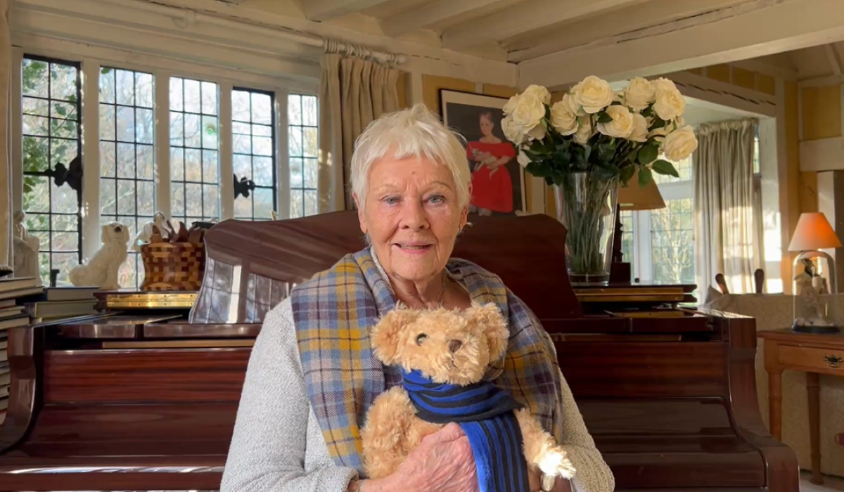 Dame Judi Dench holding a teddy bear with the College scarf. In the background, is a well lit room with a grand piano.