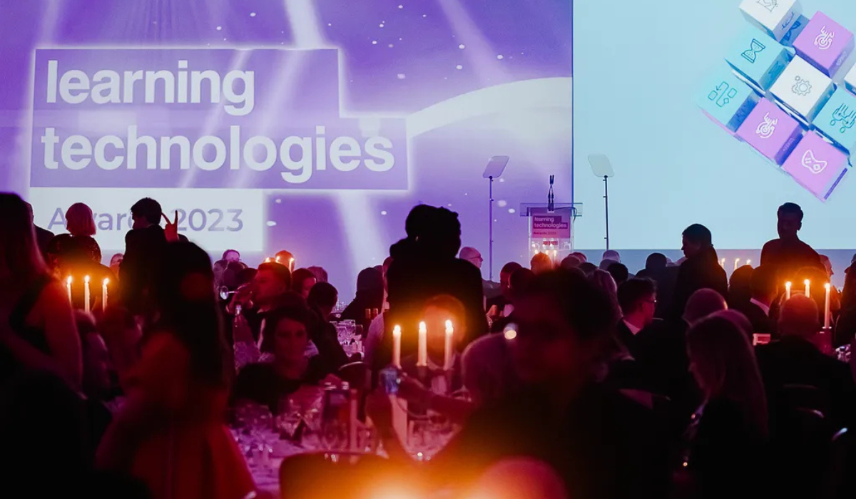 People seated at a formal candle-lit event with a stage displaying the text 'Learning Technologies Awards 2023' in the background. The stage is set up with microphones and podiums, while attendees engage in conversations around elegantly set tables.