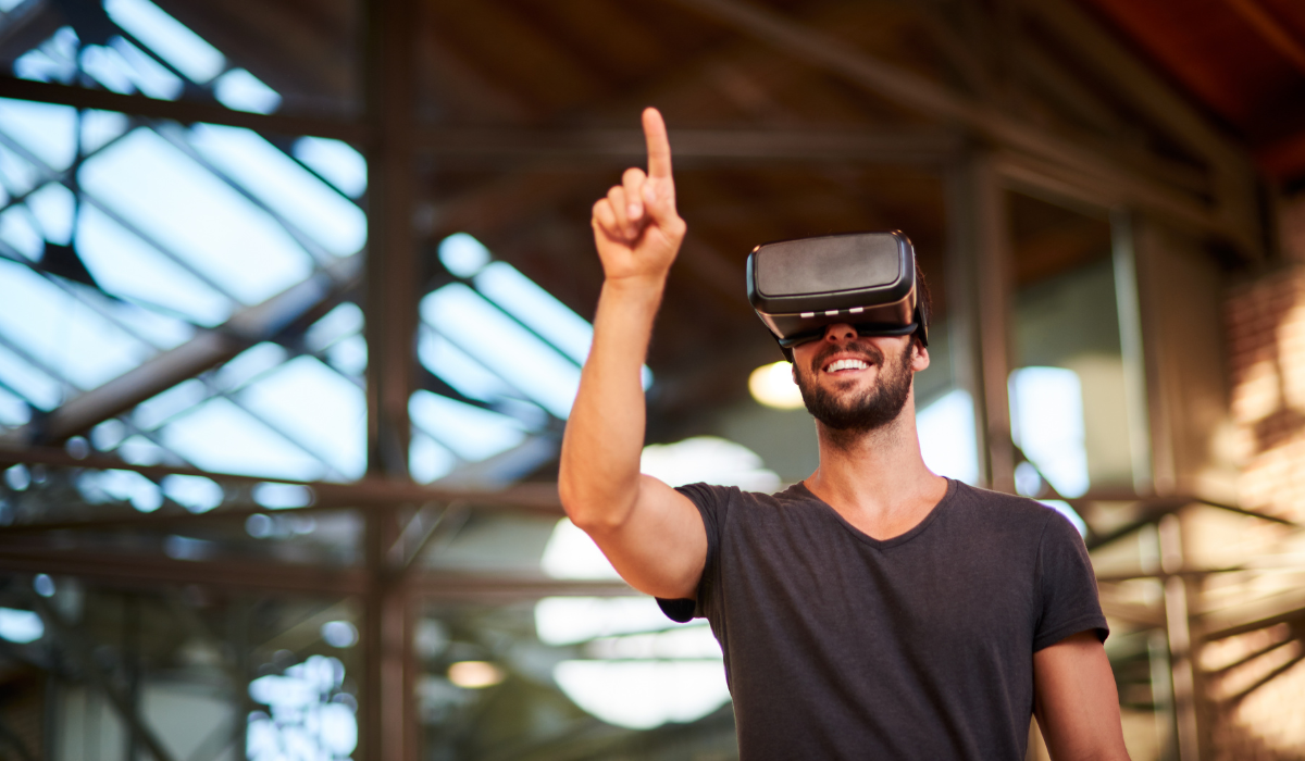 A man wearing a virtual reality headset is smiling and pointing upwards with his index finger. He is in an industrial-style indoor space with glass ceilings and metal beams visible in the background. The man appears to be interacting with a virtual environment.