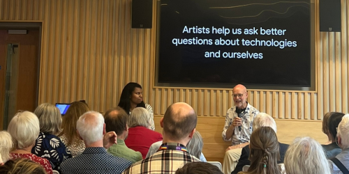 Panel event with Pamela and Alan in the cafe, in front if seated audience. The screen behind them displays a message that reads: "Artists help us ask better questions about technologies and ourselves." 