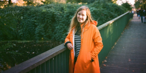Matilda posing for picture on a bridge surrounded by greenery. She is wearing an orange raincoat and stripey tshirt