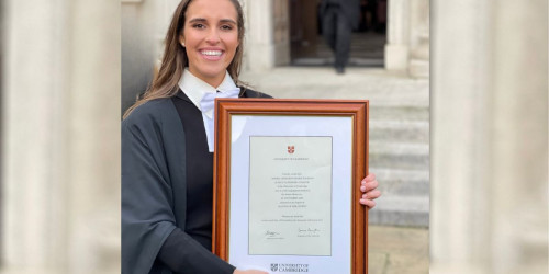 Sophia holding a framed diploma from the University of Cambridge. She is standing outside the Senate House.