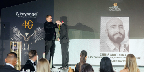 Chris is receiving a medal on stage at the awards ceremony. Behind them is a large screen displaying the name CHRIS MACDONALD and his black-and-white portrait. The text on the screen also mentions "LUCY CAVENDISH COLLEGE, UNIVERSITY OF CAMBRIDGE" and "& INNOVATION". The backdrop includes the logo for PayAngel and the event branding 40 UNDER 40 UNITED KINGDOM with an eagle motif. In the foreground, there are seated guests dressed formally, watching the stage.