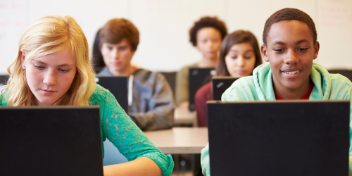 A group of students in a classroom, each working on a laptop. The focus is on two students in the foreground, one wearing a green top and the other in a light green hoodie, both engaged in their work.