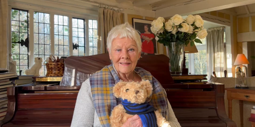 Dame Judi Dench holding a teddy bear with the College scarf. In the background, is a well lit room with a grand piano.