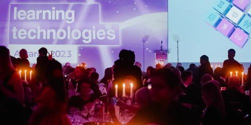 People seated at a formal candle-lit event with a stage displaying the text 'Learning Technologies Awards 2023' in the background. The stage is set up with microphones and podiums, while attendees engage in conversations around elegantly set tables.